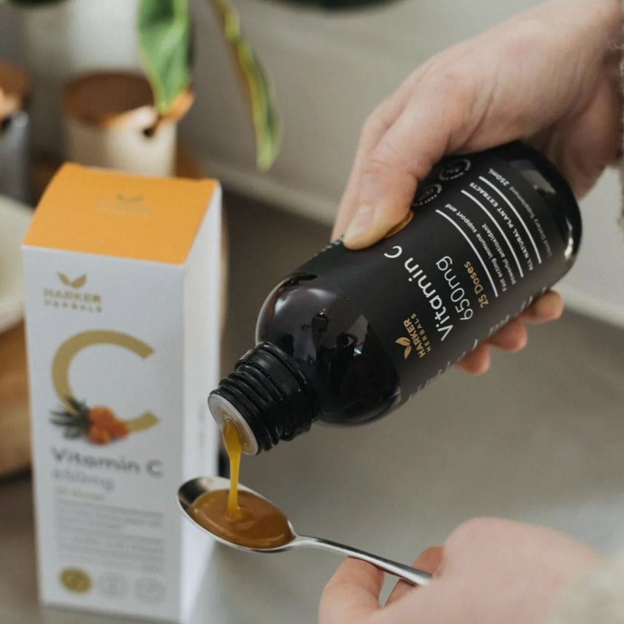 Person pouring liquid from a bottle labeled 'Vitamin C' into a spoon, with a box of the same product in the background.