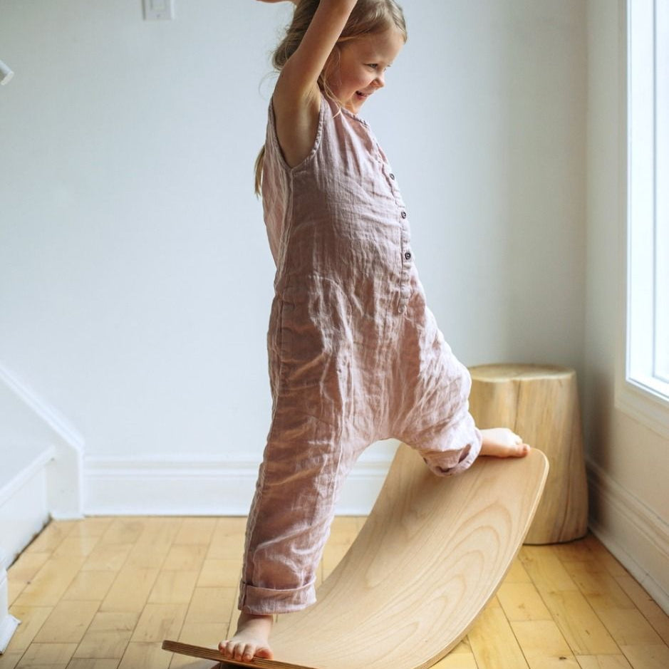 Child in pink romper standing on a wooden balance board indoors.
