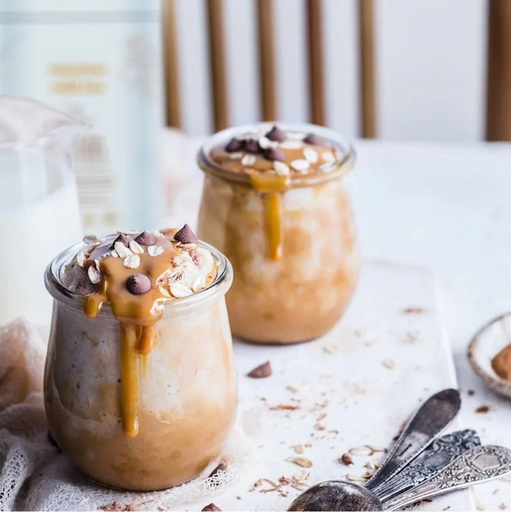 Two glass jars filled with a creamy dessert topped with chocolate chips and caramel sauce, placed on a white surface.