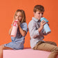 Two children sitting on a pink surface against an orange background, each holding a colorful water bottle.