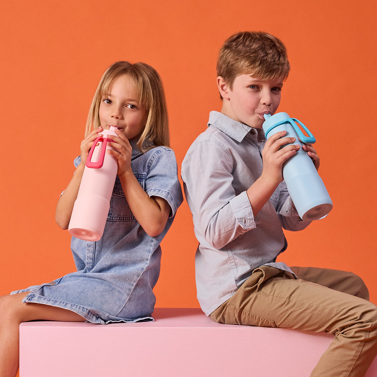 Two children sitting on a pink surface against an orange background, each holding a colorful water bottle.