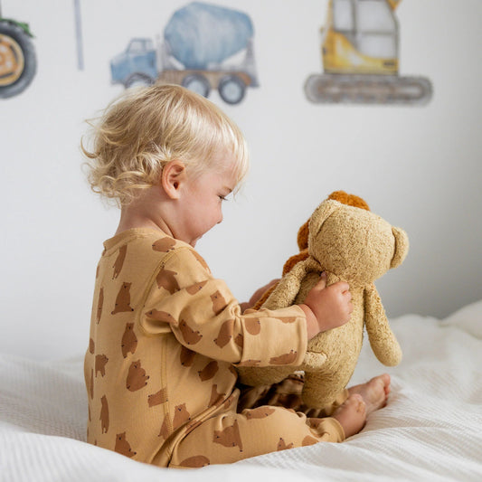 Child in a brown patterned onesie holding a teddy bear on a bed with construction-themed wall decor.