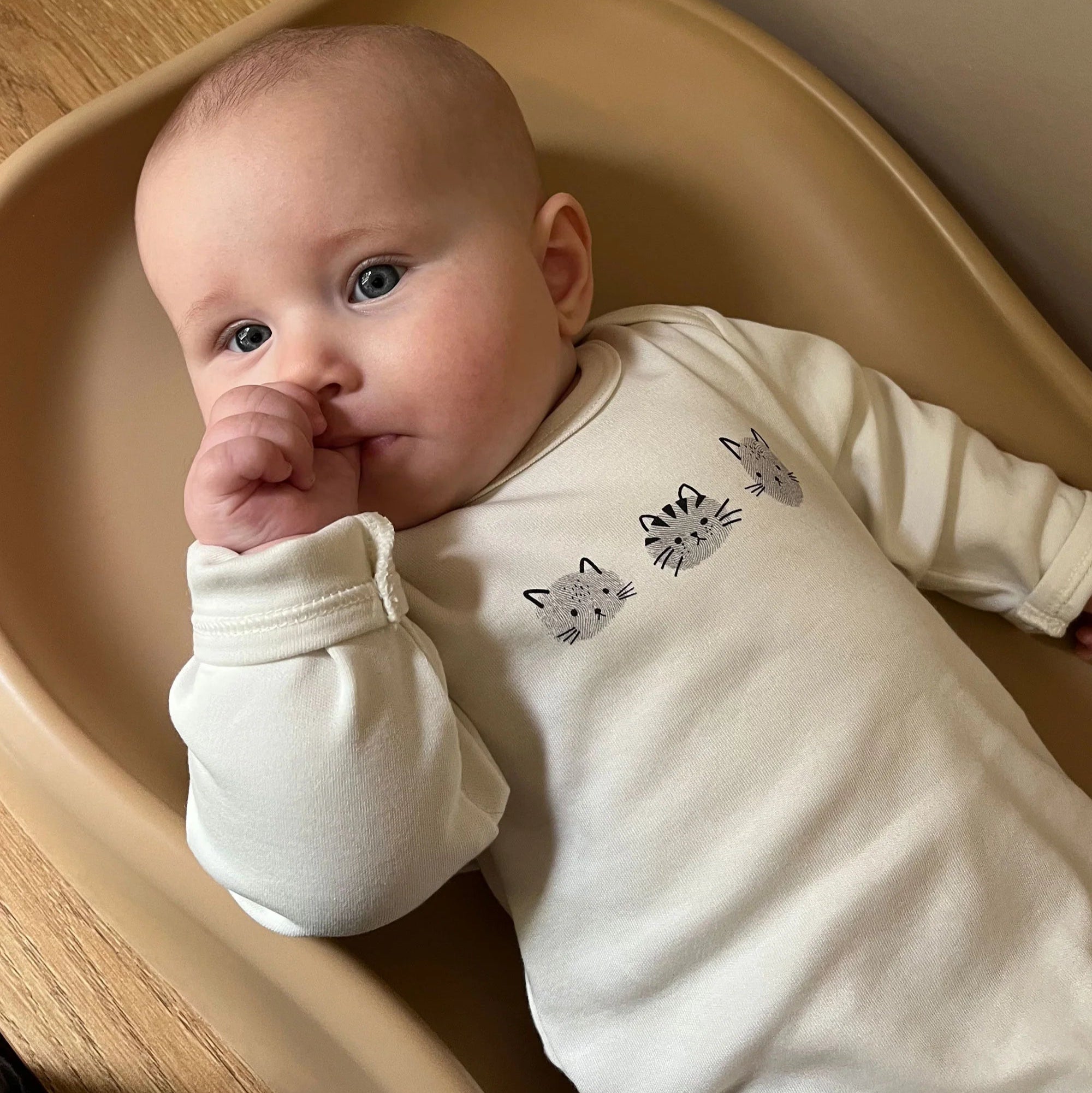 Baby lying in a beige tray wearing a white onesie with cat designs.