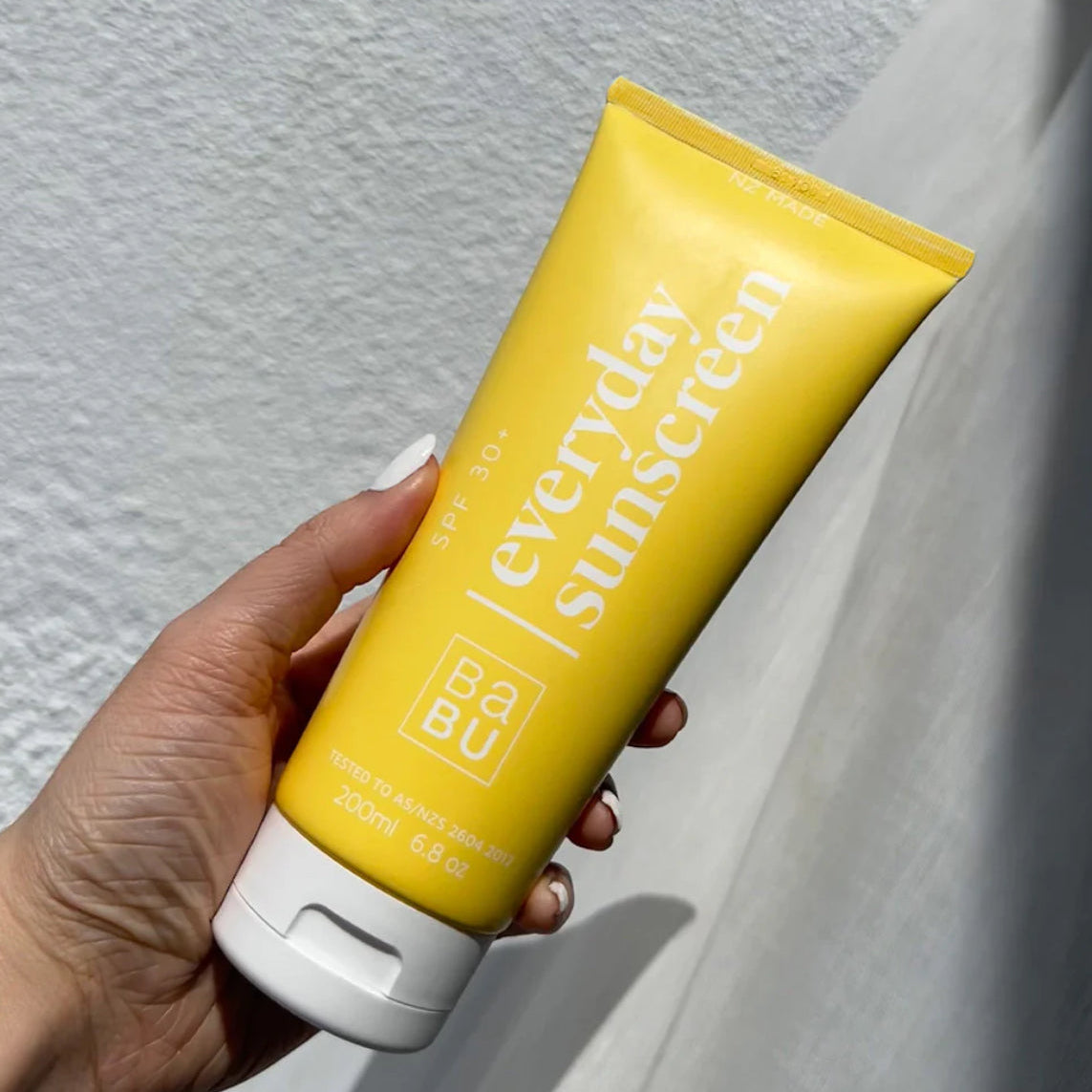 Hand holding a yellow sunscreen tube labeled 'everyday sunscreen' against a light background