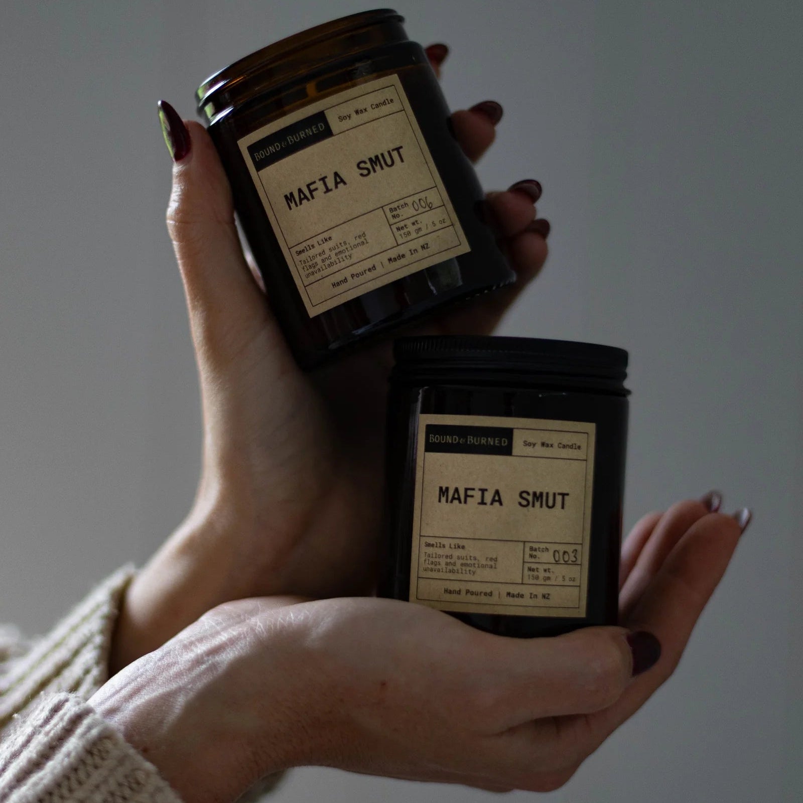 Two hands holding two 'Mafia Smut' candles against a plain background