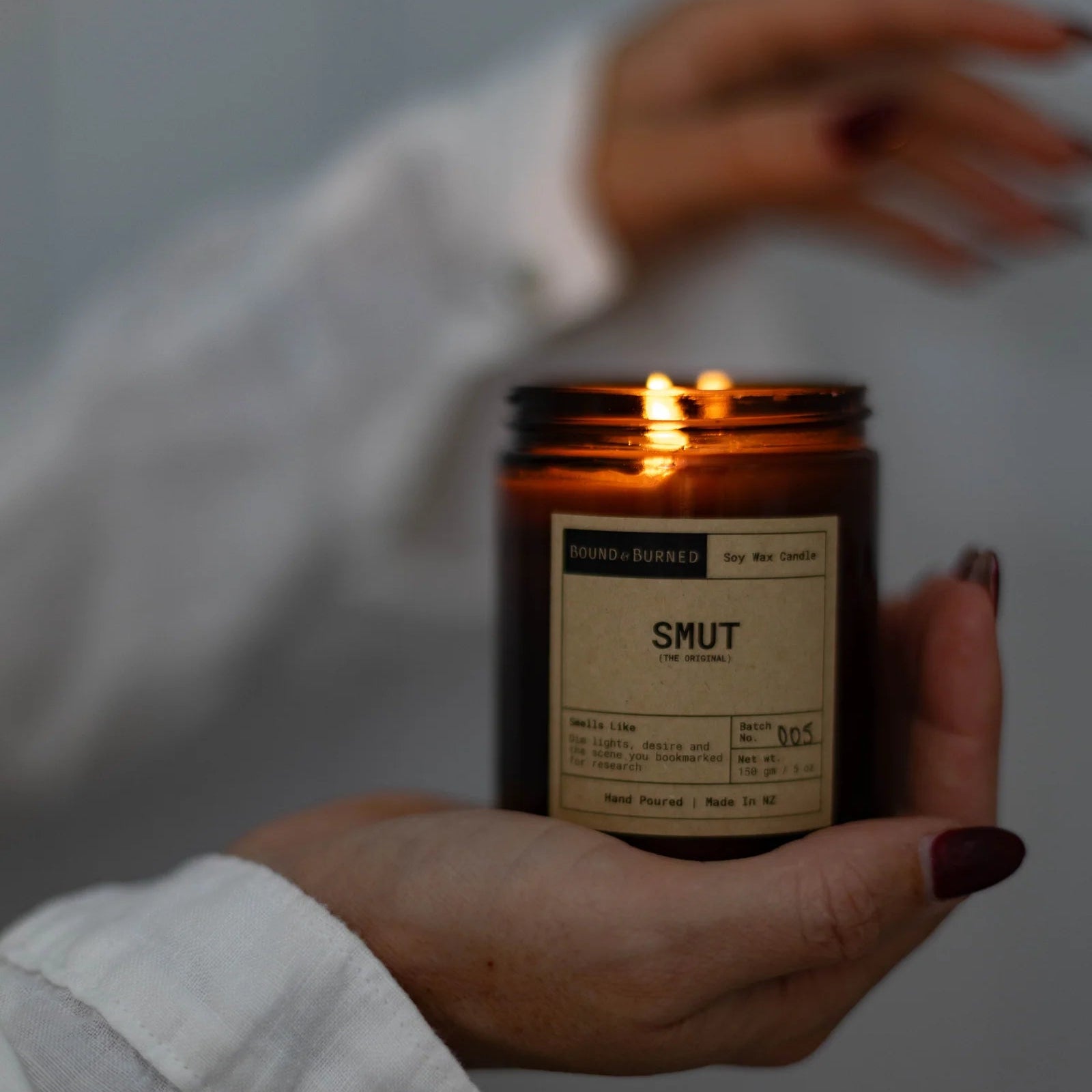 Hand holding a lit candle in an amber jar labeled 'Smut' against a neutral background