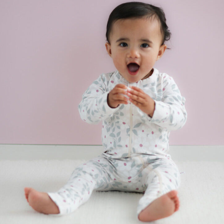 Baby sitting on a white surface wearing a white onesie with small patterns against a light pink background