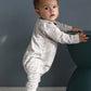 Baby in a light gray onesie standing next to a dark green stool against a gray wall.