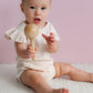 Baby in a white outfit holding a wooden rattle against a pink background