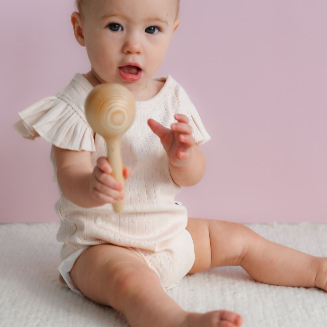 Baby in a white outfit holding a wooden rattle against a pink background
