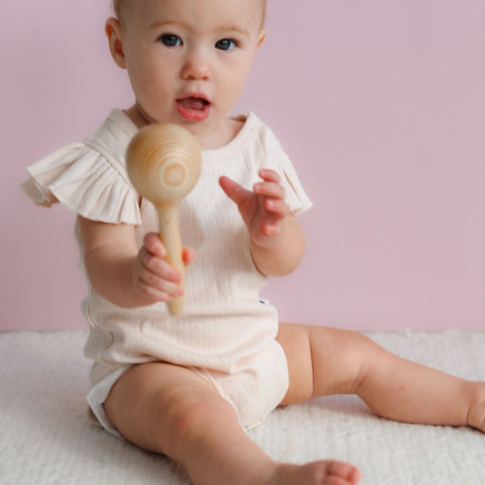 Baby in a white outfit holding a wooden rattle against a pink background