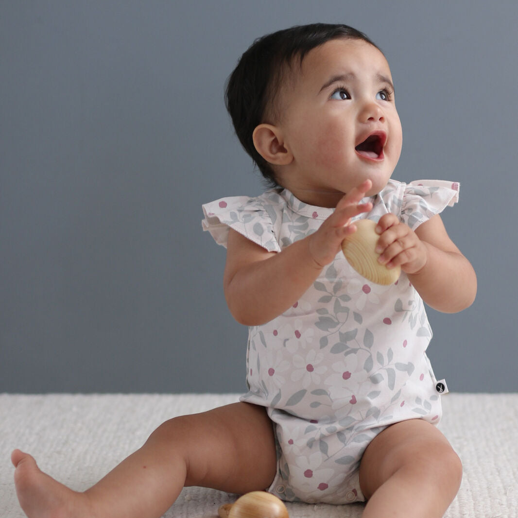 Baby sitting on a white surface with wooden toys, wearing a patterned onesie against a gray background.
