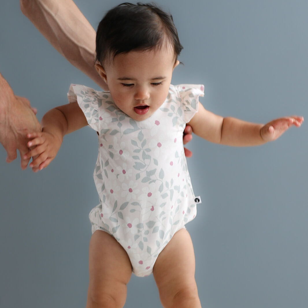 Baby in a white onesie with ruffled sleeves standing on a light surface against a gray background
