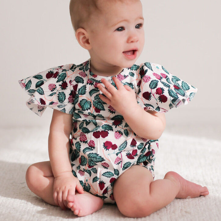 Baby wearing a floral romper sitting on a light-colored blanket.