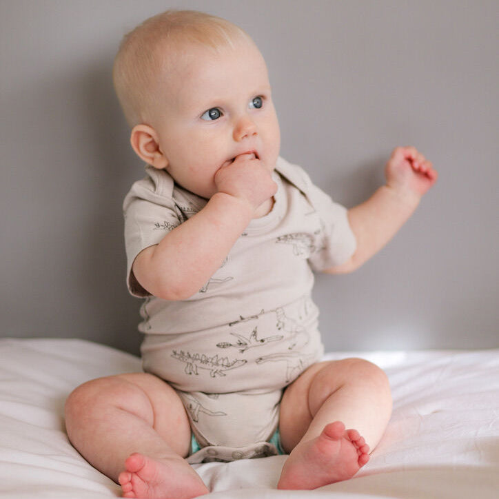 Baby sitting on a bed wearing a light-colored onesie.