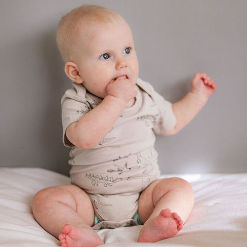 Baby sitting on a bed wearing a light-colored onesie.