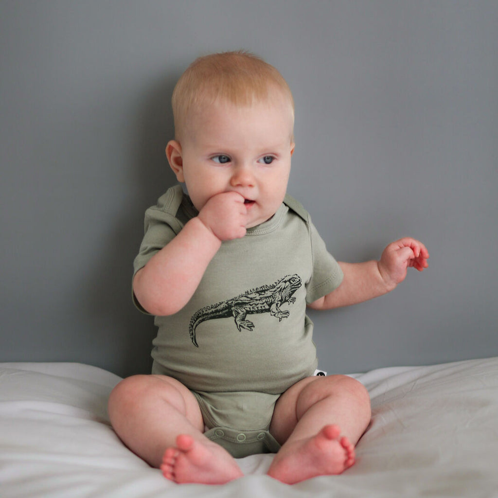 Baby wearing a green onesie with a lizard design, sitting on a white surface against a gray background.