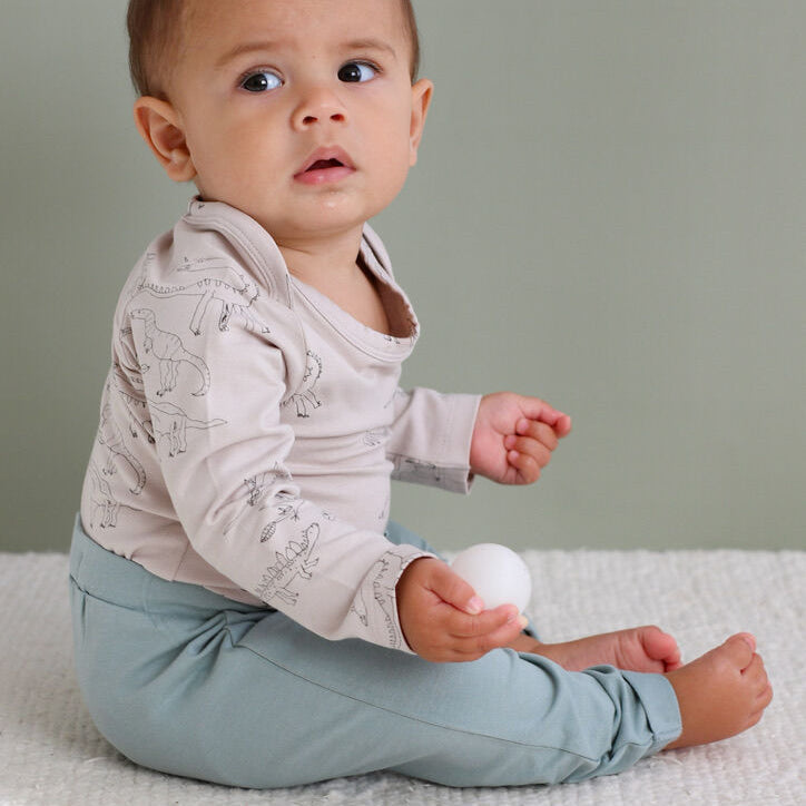 Baby sitting on a textured surface wearing a light grey top with dinosaurs and light blue pants.