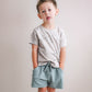 Young child wearing a light gray shirt and green shorts against a plain background