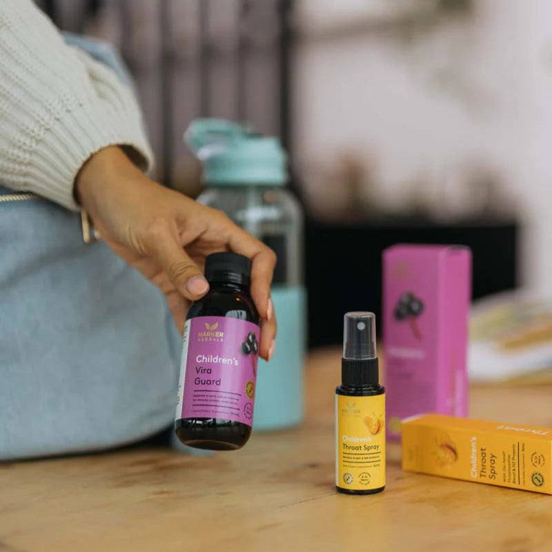 Hand holding a bottle of vira guard with a blurred background showing a water bottle and throat spray.