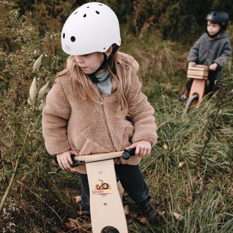 Two children riding balance bikes in a natural setting with grass and plants.