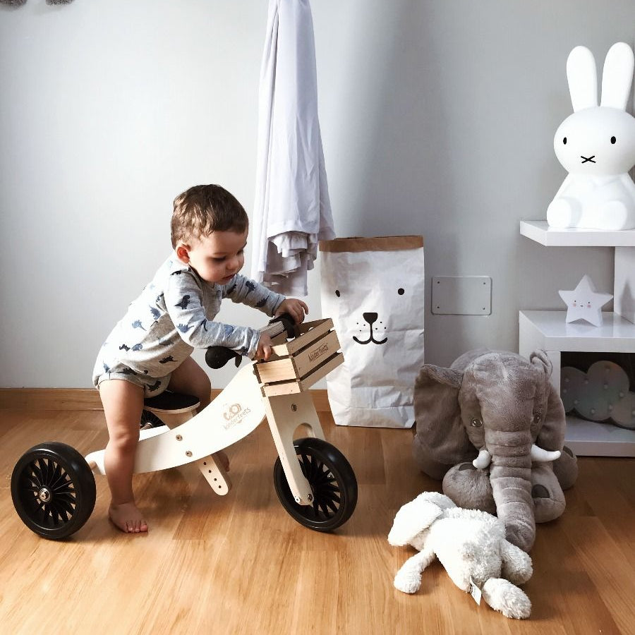 Child playing with a wooden bike in a room with toys and a curtain.
