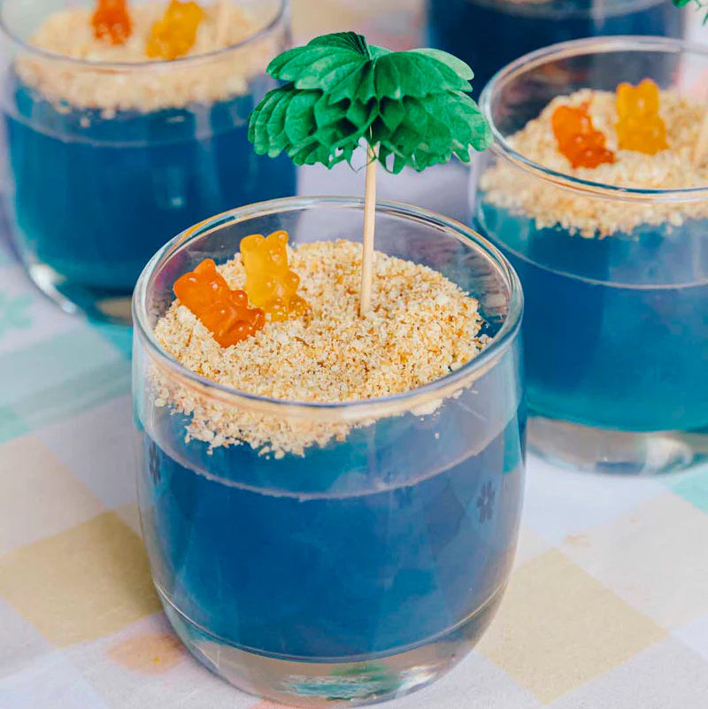 Glass cups with blue jelly, gummy bears, and sand-like topping on a checkered tablecloth.