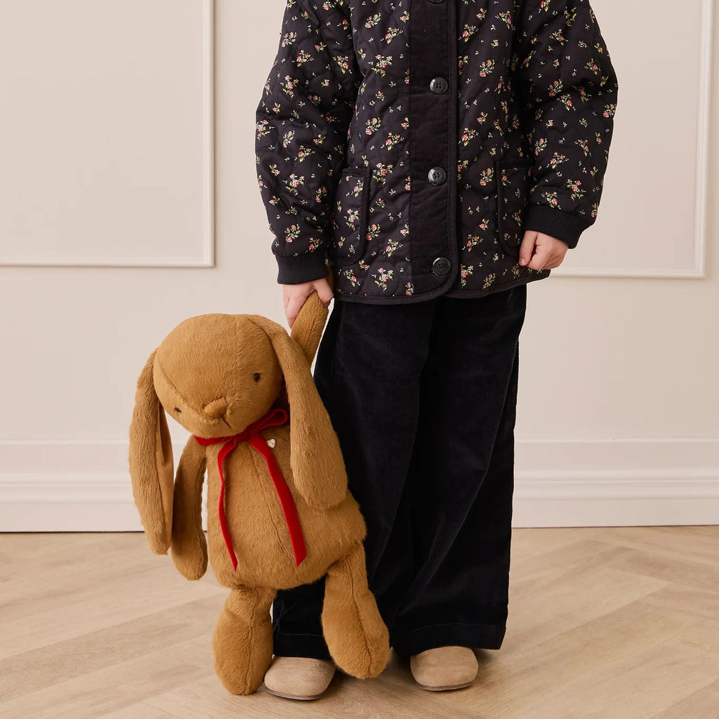 Child wearing a dark floral jacket holding a plush bunny toy against a light-colored wall.