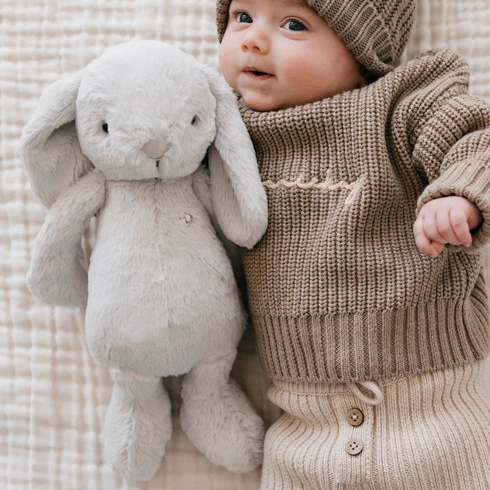 Baby in knitted outfit holding a plush toy on a textured blanket
