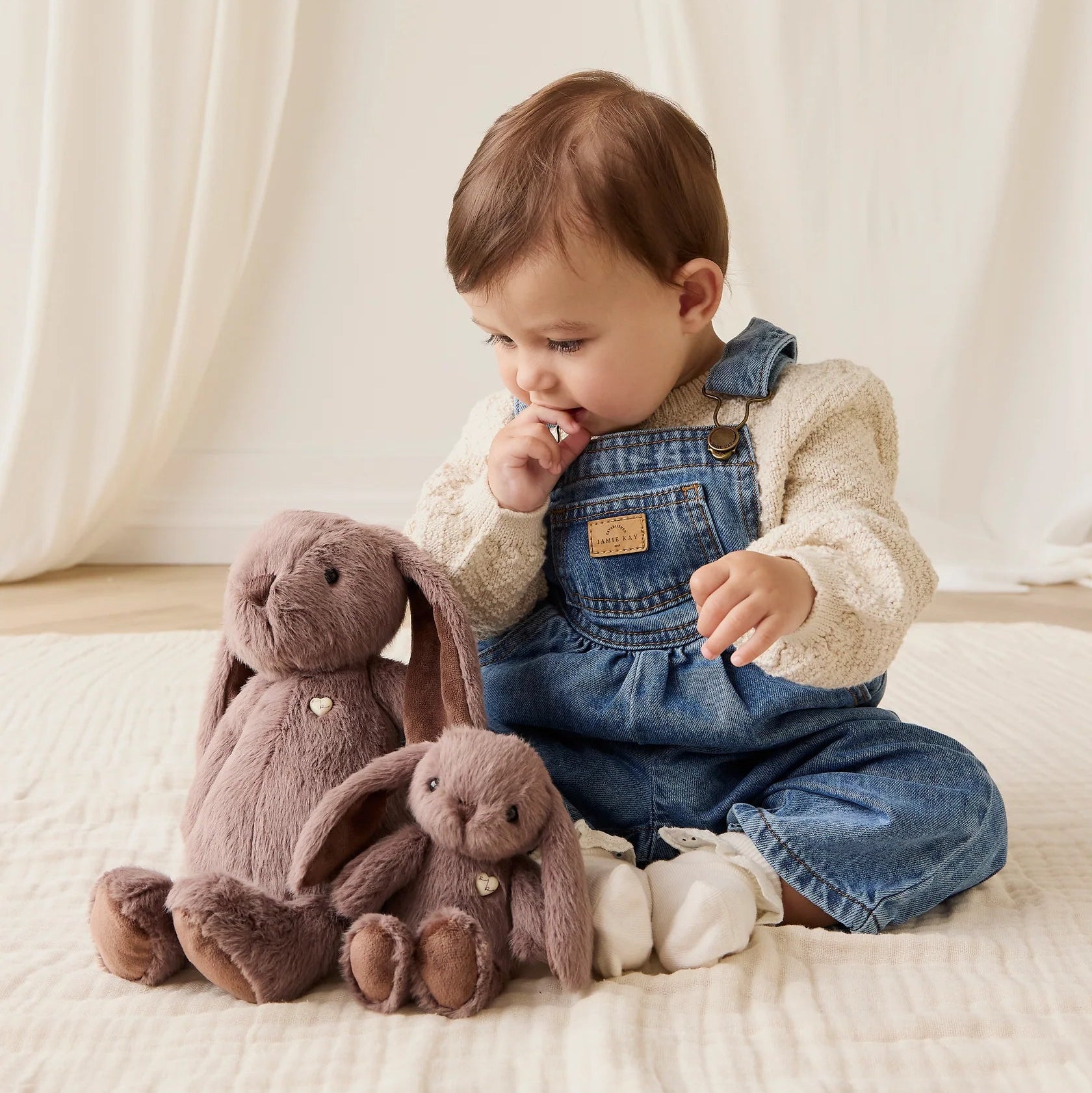 Child sitting on a bed with two plush bunny toys, wearing denim overalls.